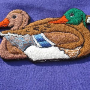 May include: Embroidered duck patch featuring a brown body, green head, and orange beak. The duck has blue and white accents on its wings and is set against a purple background. The patch is likely for clothing or accessories.