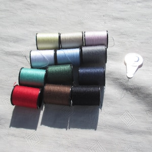 May include: A collection of 12 spools of thread in various colors, including red, green, blue, black, and white. The spools are arranged in a stack and are ready for sewing.