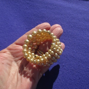 May include: A vintage bracelet featuring a circular design made of cream-colored faux pearls. The bracelet has a gold-tone metal clasp. The pearls are arranged in multiple rows, creating a textured and dimensional look. The bracelet is held in a hand against a purple background.
