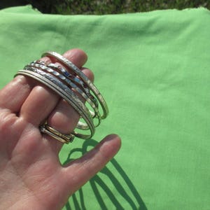 May include: A set of four silver-toned metal bangles and one gold-toned ring. The bangles feature varied textures, including a hammered finish, a smooth surface, and intricate engraved patterns. The ring is a simple, thin gold-toned band.