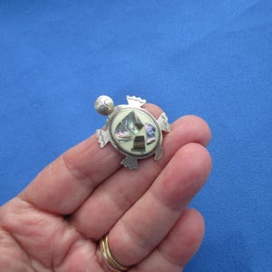 May include: A silver-toned turtle brooch featuring an oval shell inlay composed of iridescent abalone shell fragments and smooth cream-colored segments. The metal body has a polished finish with scalloped edges on the flippers and a rounded head.