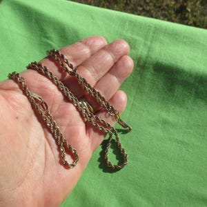 May include: A gold-toned rope chain necklace. The chain is held in a hand against a green background. The necklace has a clasp closure and a twisted rope design. The chain is approximately 20 inches long.