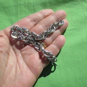 May include: A silver-colored chain bracelet with an oval link design. The bracelet is held in a hand against a green background. The links are interlocked, creating a textured appearance. The bracelet is likely made of metal.