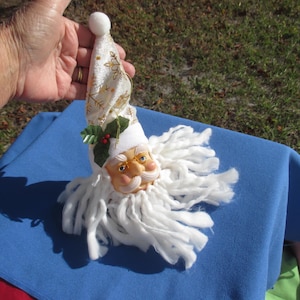 May include: A Santa Claus Christmas ornament with a white beard and a cream-colored face. The hat is cream-colored with gold snowflake designs. The ornament has a white pom-pom at the top of the hat and a sprig of holly.
