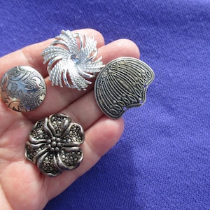 May include: A collection of five vintage silver-toned buttons with intricate designs. The buttons feature floral, swirl, and shell-like patterns. The buttons are displayed on a purple background.