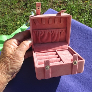 May include: Open, pink jewelry box with a soft, velvety interior. The box has multiple compartments for rings, earrings, and other small items. It features a clasp closure and a handle on the top. The box is held open by a hand.