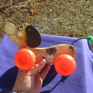 May include: A vintage wooden pull-along toy dog with orange wheels. The dog has a brown ear, a black spot, and a green ball at the end of its spring tail. The toy is tan and brown with black accents.