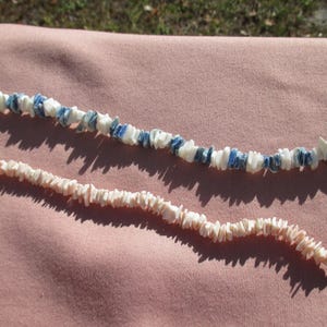 May include: Two necklaces made of small, irregular shell pieces. One necklace features white and blue shells, while the other is made of off-white shells. Both necklaces are on a pink surface.