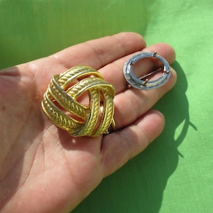 May include: Two vintage brooches. One is a gold-toned, woven knot design. The other is a silver-toned, circular brooch with a pin closure. Both are displayed on a green background.