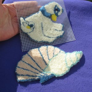 May include: Two handmade, textured fiber art pieces: a white bird with blue and yellow accents, and a fan-shaped design with blue, beige, and white colors. The pieces are on a white grid, set against a purple background.