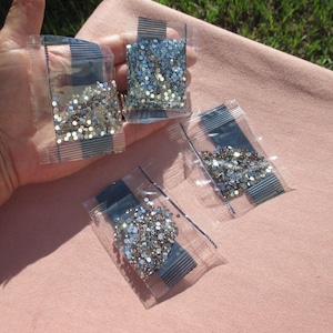 May include: Four small, clear plastic bags filled with sparkling, iridescent rhinestones. The bags are sealed with a dark blue strip and are held against a pink fabric background. The rhinestones vary in color, including silver and light blue.