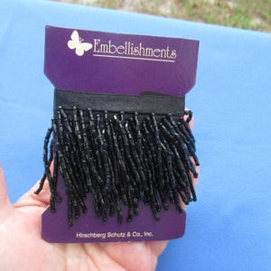 May include: A spool of black beaded fringe trim wrapped around a purple cardboard card. The card features a white butterfly icon, the text Embellishments, and the text Hirschberg Schutz & Co., Inc. The fringe consists of numerous thin black beads.