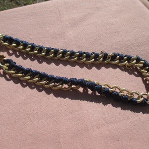 May include: A gold-tone chain necklace with navy blue woven accents. The necklace features a chunky chain design with a textured finish. The clasp is also gold-toned. The necklace is displayed on a pink surface.