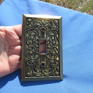 May include: Ornate brass-colored light switch cover with a rectangular opening and intricate floral and scrollwork design. The cover has a rectangular shape with a textured border and is held against a blue background.