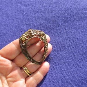 May include: Ornate antique brass brooch with an openwork design. The brooch has a detailed, filigree-style frame with a central open oval. The metal has a warm, aged patina, suggesting vintage craftsmanship. The brooch is held in a hand against a purple background.