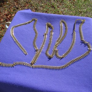 May include: Several gold-colored metal chains with a repeating leaf-like design are arranged on a purple cloth. The chains are laid out in a casual, overlapping pattern, catching the light. The background includes grass and other fabrics.