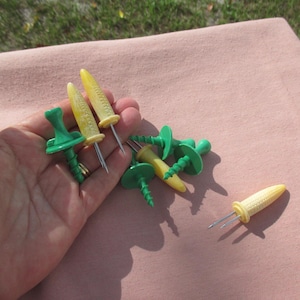 May include: A collection of corn-on-the-cob holders featuring yellow, textured corn-shaped handles and green, solid-colored handles. Each holder is equipped with two sharp metal prongs designed for securing corn, with some pieces showing a spiral design.