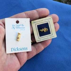 May include: Two small cards with gold pins. The card on the left has a gold pin of two hands clasped in prayer. The card on the right has a gold pin of a cross. The card on the left says "You Shall Be My Witnesses ACTS 1:8" and "Dicksons". The card on the right says "FRANCISCAN FATHERS" and "NEW CANAAN, CONN."