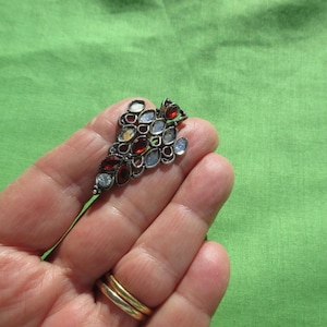 May include: A silver-toned brooch with a cluster of colorful gemstones. The brooch features red, clear, and light blue stones set in an ornate, leaf-like design. The brooch is held in a hand against a green background.