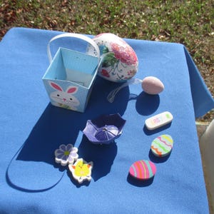May include: An assortment of Easter-themed decorations on a blue surface. Includes a blue polka-dot basket with a bunny, decorated eggs, a purple umbrella, and a small eraser with the word "EASTER".
