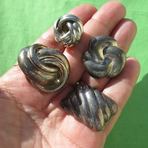 May include: Four silver-toned vintage earrings in various designs, including knot and swirl shapes. The earrings are displayed on a hand against a green background, showcasing their metallic sheen and intricate details.