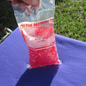 May include: A clear plastic bag containing 150 red ruby sunburst beads from The Beadery. The packaging features the text 950B 10 mm 150 SUNBURST, 150 Perles SUN, 150 SUNBURST, 150 Cuentas SUNBURST, and 017 RUBY, with a Made in U.S.A. label.