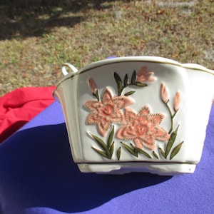 May include: A white ceramic planter with a floral design. The planter features embossed peach-colored lilies with green leaves and stems. The planter has a scalloped rim and small handles.