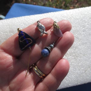 May include: A collection of small, decorative charms and pins. Includes a silver fish charm, a blue and gold triangular pin, a purple teardrop-shaped charm with gold accents, and a blue beaded charm. The items are held in a hand.