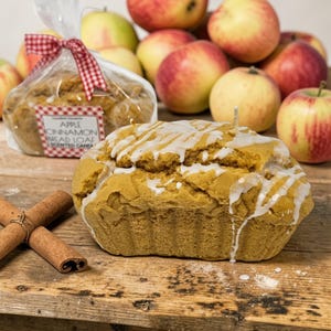 May include: A loaf-shaped candle with white icing, cinnamon sticks, and a bag of bread loaf candles. The bag is tied with a red and white checkered ribbon. Apples are in the background.