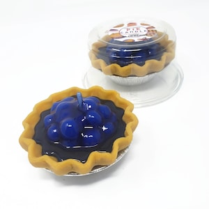 Puede incluir: Dos velas con forma de tarta de ar&aacute;ndanos. Las velas tienen un relleno azul oscuro con una mecha de vela azul claro. La corteza es de color marr&oacute;n claro y se encuentra en una lata plateada. Una vela est&aacute; en un recipiente de pl&aacute;stico transparente con el texto "PIE CANDLE BLUEBERRY".