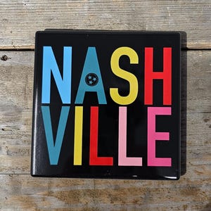 May include: Square black sign with the word "NASHVILLE" in large, colorful letters. The letters are blue, yellow, red, and pink. A small emblem with three stars is above the "V" in "NASHVILLE".