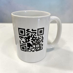 May include: White ceramic mug featuring a large black QR code. The mug has a curved handle and a simple design. The background is a light blue and white. The mug is suitable for hot beverages.