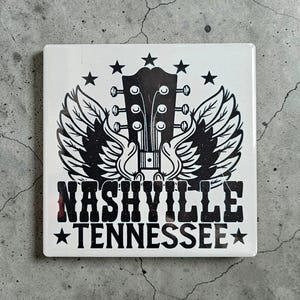 May include: Square white coaster with a black and white design. The design features a guitar headstock with wings, stars, and the words "Nashville Tennessee". A decorative item for music lovers.