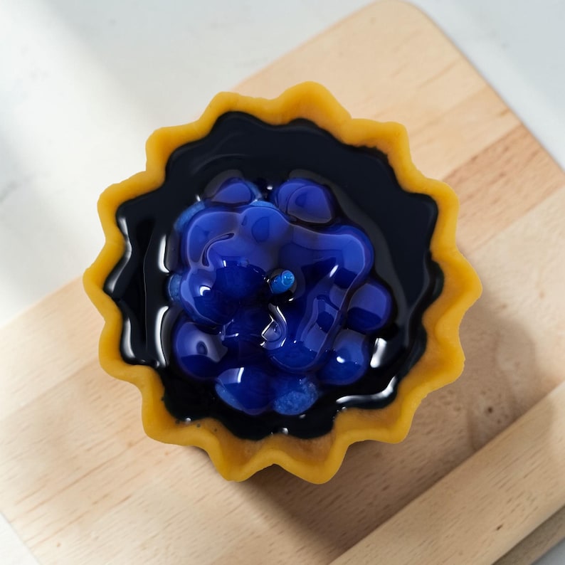 Puede incluir: Primer plano de una tarta en miniatura con una corteza amarilla y festoneada. La tarta est&aacute; rellena de gelatina azul oscuro y cubierta con caramelos esf&eacute;ricos azules, que se asemejan a ar&aacute;ndanos. La tarta est&aacute; sobre una superficie de madera marr&oacute;n claro.