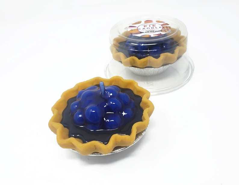 Puede incluir: Dos velas con forma de tarta de ar&aacute;ndanos. Las velas tienen un relleno azul oscuro con una mecha de vela azul claro. La corteza es de color marr&oacute;n claro y se encuentra en una lata plateada. Una vela est&aacute; en un recipiente de pl&aacute;stico transparente con el texto "PIE CANDLE BLUEBERRY".