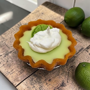 May include: A lime key pie candle with a light green filling, topped with white whipped cream and a lime slice. The candle sits in a brown, fluted crust on a wooden surface, with fresh limes nearby.