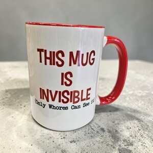 May include: White ceramic mug with a red interior and handle. The mug features the text "THIS MUG IS INVISIBLE (Only Whores Can See It)" in red lettering. The mug is on a gray surface.