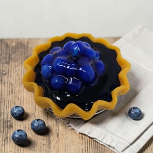 Puede incluir: Una vela decorativa con forma de tarta de ar&aacute;ndanos. La vela presenta una corteza amarilla, un relleno azul oscuro y un grupo de ar&aacute;ndanos en la parte superior. Varios ar&aacute;ndanos frescos est&aacute;n esparcidos alrededor de la tarta sobre una superficie de madera.