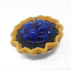 Puede incluir: Una peque&ntilde;a tarta de ar&aacute;ndanos con una corteza dorada y un relleno azul oscuro brillante. La tarta est&aacute; en un peque&ntilde;o recipiente de papel de aluminio plateado. Un solo tallo azul se eleva desde la parte superior.