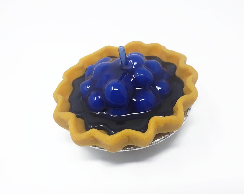 Puede incluir: Una peque&ntilde;a tarta de ar&aacute;ndanos con una corteza dorada y un relleno azul oscuro brillante. La tarta est&aacute; en un peque&ntilde;o recipiente de papel de aluminio plateado. Un solo tallo azul se eleva desde la parte superior.
