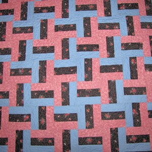 May include: A handmade quilt with a geometric pattern. The design features interlocking blocks in light blue, pink, and black with floral accents. The quilt has a black floral border and a blue binding.