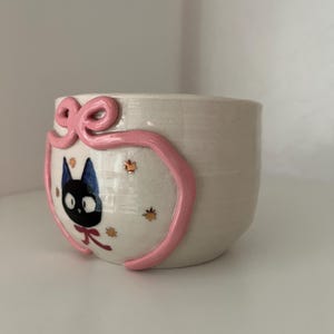 May include: A small, off-white ceramic cup with a pink ribbon design. The cup features a cartoon black cat with a blue head and a red bow. Gold and orange leaf accents are also present.