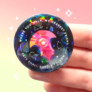 Op de afbeelding: Een ronde, holografische knoop met de tekst "WHERE YOU WALK, MY DEAREST FRIEND, FATE SHALL SURELY FOLLOW." De knoop heeft een geometrisch ontwerp met een centrale roze edelsteen en sterrenbeeldpatronen.