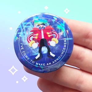 Peut inclure: Un bouton rond bleu avec un effet holographique représentant un personnage de dessin animé en veste rouge, pantalon noir et gants jaunes. Le texte "I'VE COME TO MAKE AN ANNOUNCEMENT" est imprimé sur le bord.