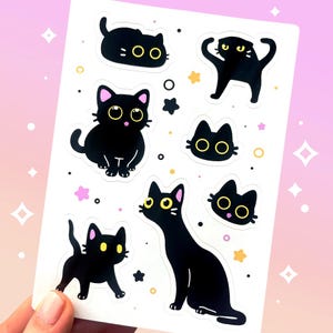 May include: A sheet of stickers featuring various black cat illustrations with yellow eyes and pink accents. The cats are in different poses, surrounded by stars and dots. The background has a pink and white gradient.