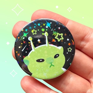 Puede incluir: Un botón redondo con un alienígena verde con antenas y una expresión de mal humor. El fondo negro está decorado con estrellas y destellos de colores. El botón se sostiene en una mano sobre un fondo verde claro.
