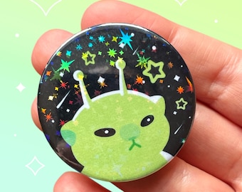 May include: A round button featuring a green alien with antennae and a grumpy expression. The background is black with colorful stars and sparkles. The button is held in a hand against a light green background.