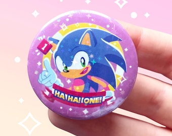 May include: A round button featuring Sonic the Hedgehog with a thumbs-up gesture. The button has a purple background with the text "HA! HA! ONE!" and a number "1" on it. The design includes stars and a yellow and white arc.
