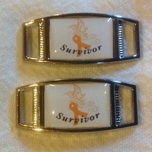 May include: Two silver shoe charms with a white background and a gold ribbon with a butterfly design. The charms say "Survivor" in black text.