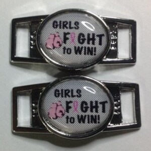Puede incluir: Dos abalorios de zapatos plateados con fondo blanco y un guante de boxeo rosa con una cinta rosa. Los abalorios dicen "Girls Fight to Win!"